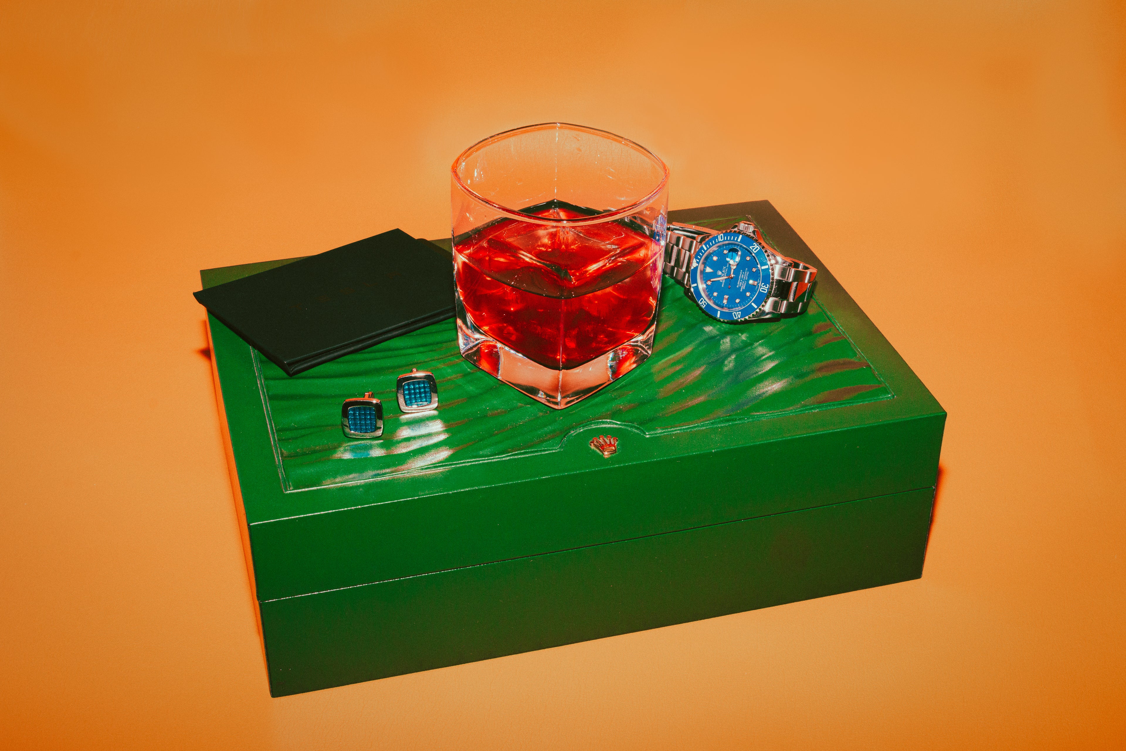 Luxurious setup featuring a glass of non-alcoholic spirit on a green Rolex box alongside a watch and cufflinks, highlighting botanical drinks and plant-based alcohol alternatives in a stylish lifestyle scene.